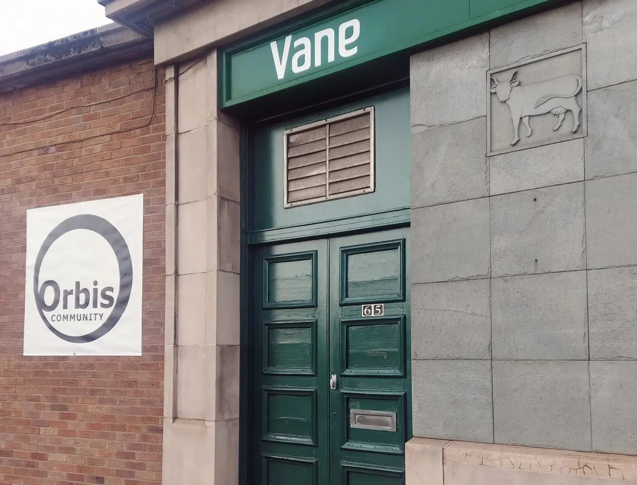 This is a photograph, so I will provide alt text:

Green double doors numbered 65 with 'Vane' signage above and 'Orbis Community' logo on adjacent brick wall.