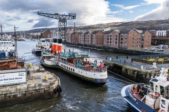 Historic paddle steamer moored at busy riverside dock with industrial crane, other vessels and waterfront buildings in background.