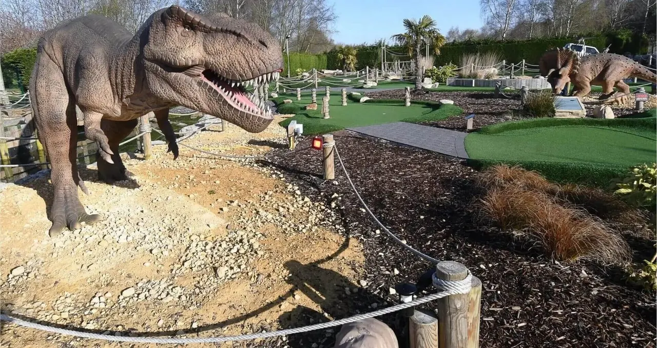 Dinosaur sculptures at a mini golf course with a large T-Rex in the foreground and smaller dinosaurs visible across the themed landscape.