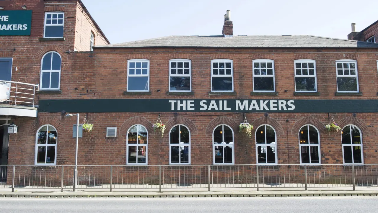 Red brick building with "The Sail Makers" signage, arched windows with hanging baskets, and metal railings across the waterfront frontage.