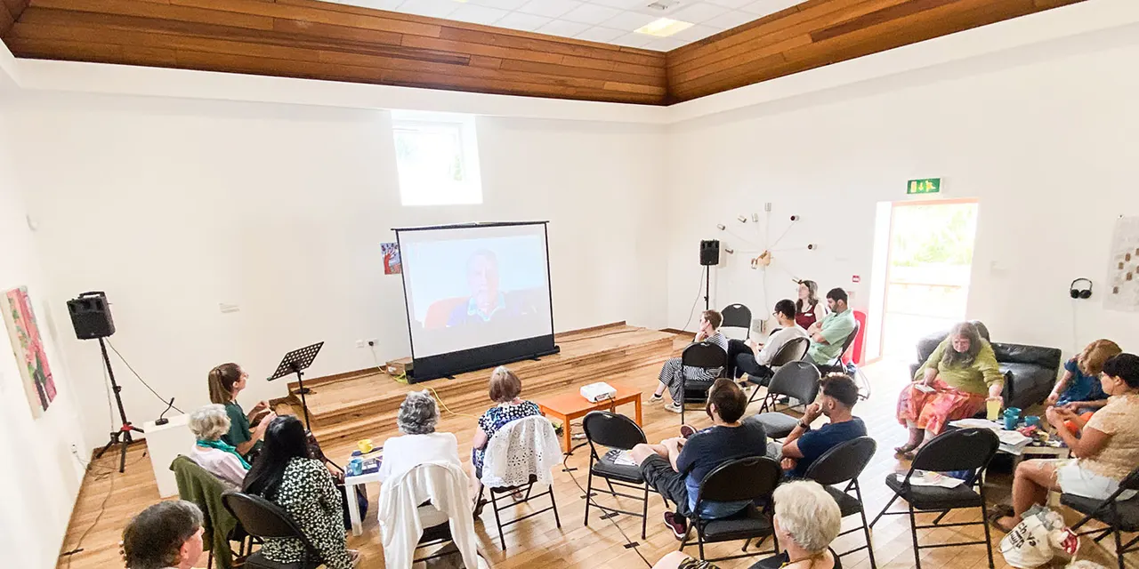 A community gathering in a wooden-beamed room where diverse adults of varying ages watch a presentation on a large screen.