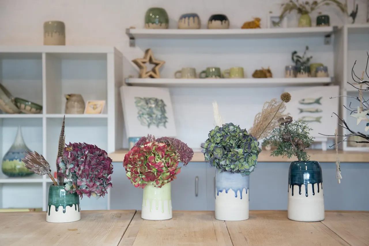 Four ceramic vases with dried flowers and hydrangeas arranged on a wooden table against white shelving displaying decorative items.