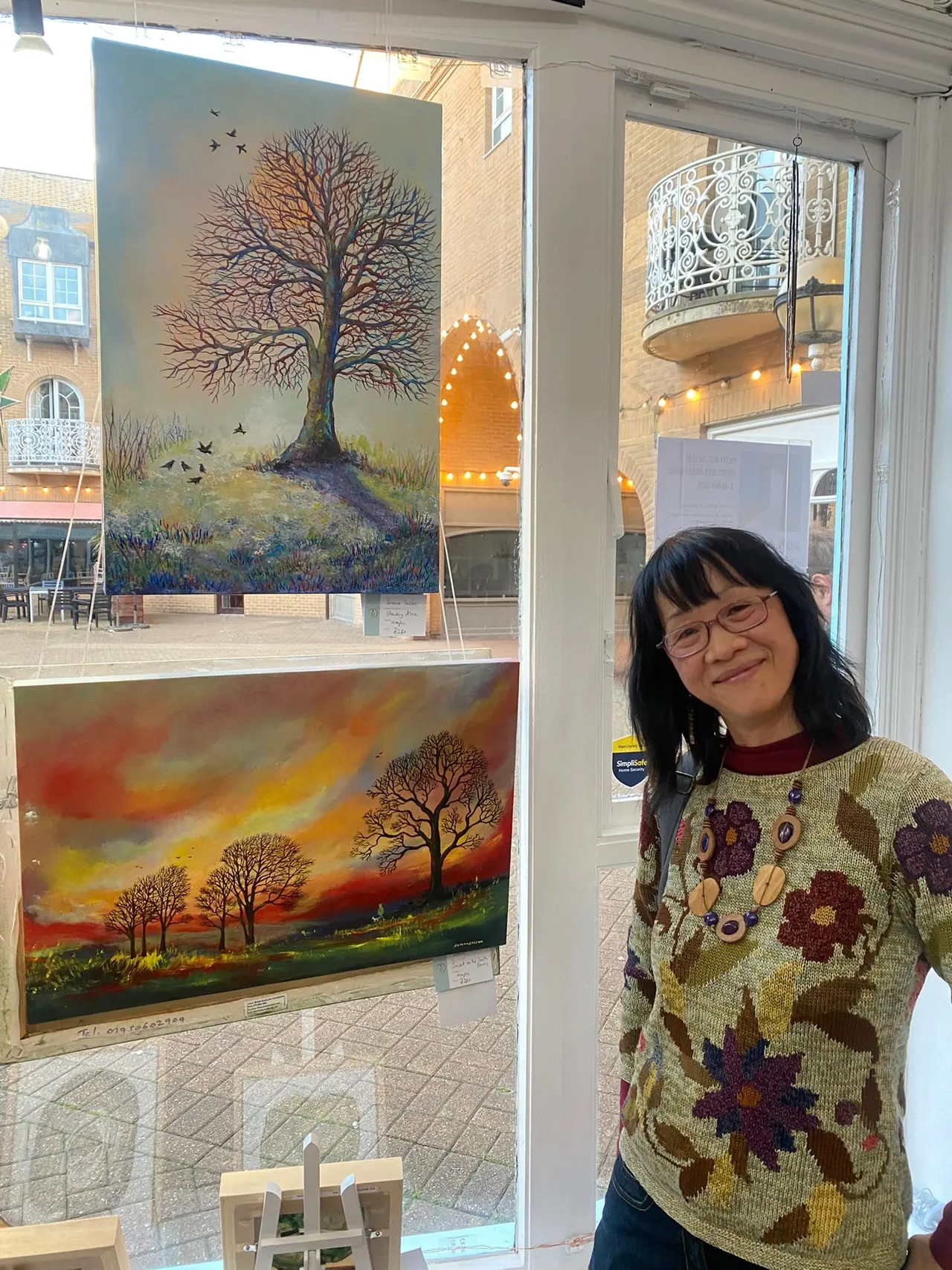 A woman wearing glasses and a patterned jumper smiles at the camera inside a gallery space displaying landscape paintings featuring bare trees.