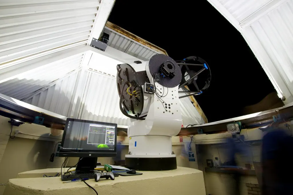 Interior of an observatory with a large telescope mounted on a white structure, control monitor visible below, opening to night sky.