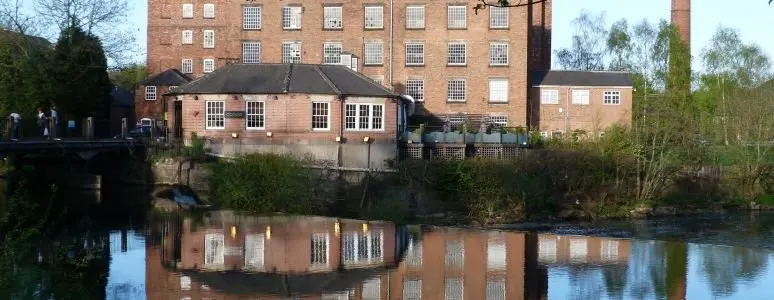Historic brick mill buildings reflected in still water alongside a river.