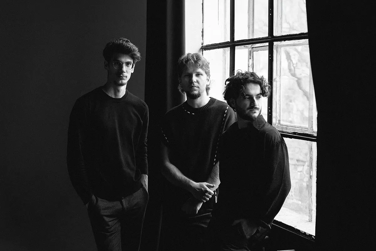 Three men stand together in a dark room beside a large latticed window in black and white portrait photograph.