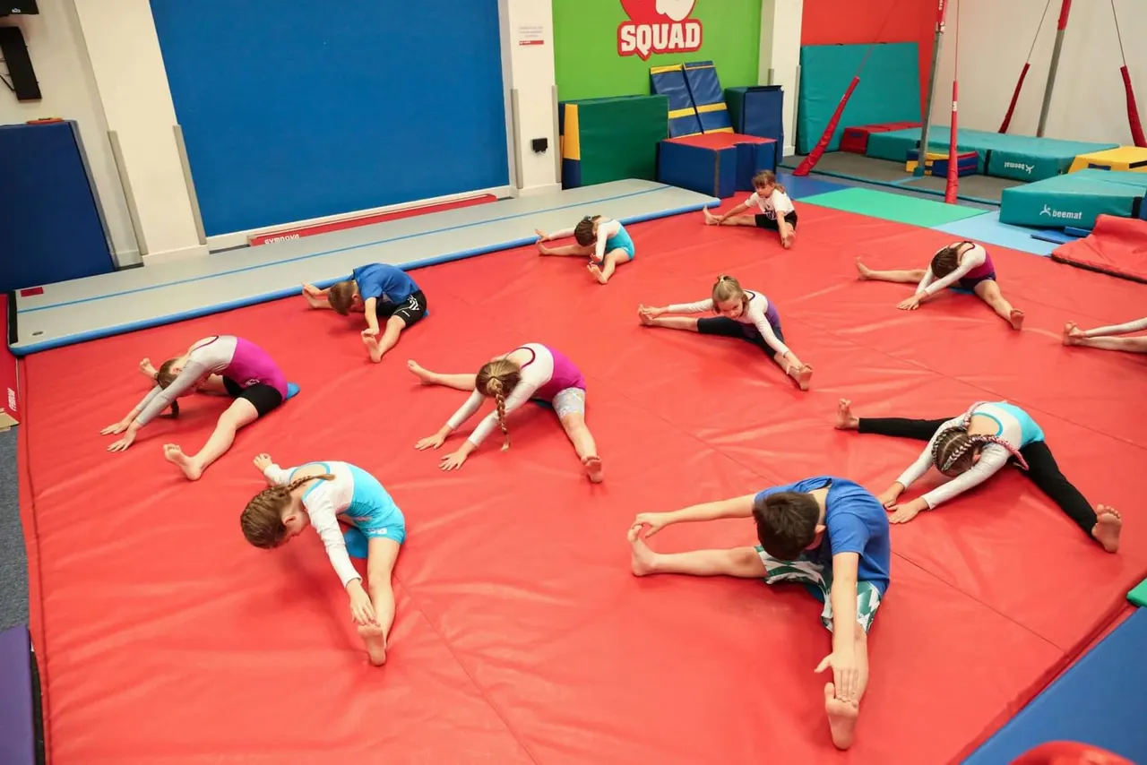 Children performing stretching exercises on a red gymnastics mat during a class at an indoor gymnastics facility.
