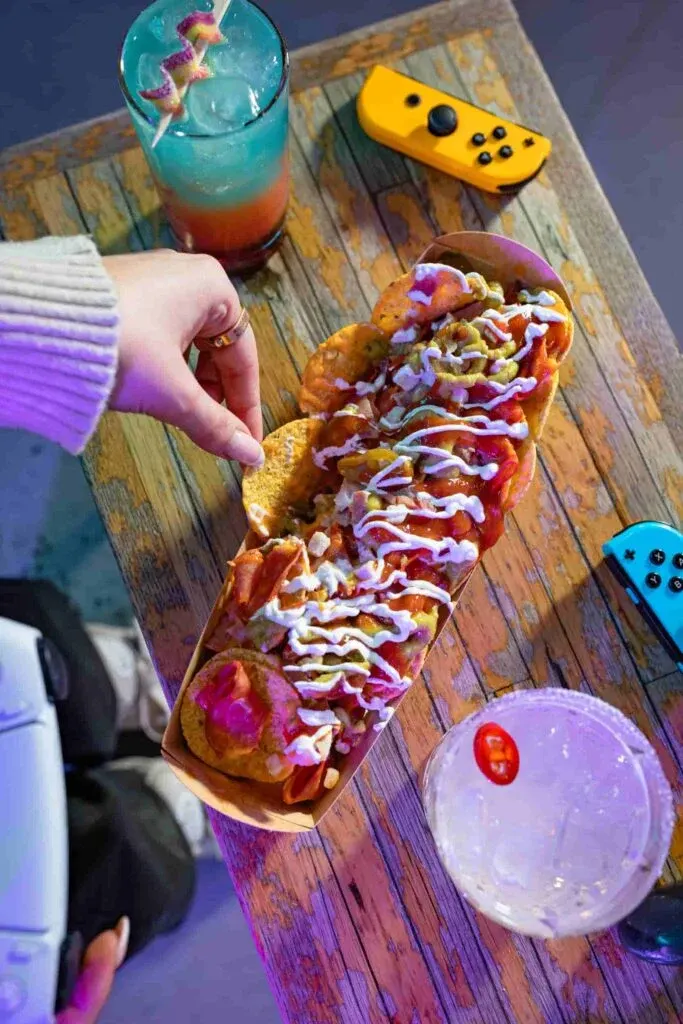 Person holding a colourful hot dog with toppings beside a turquoise cocktail and gaming controller on a wooden table.