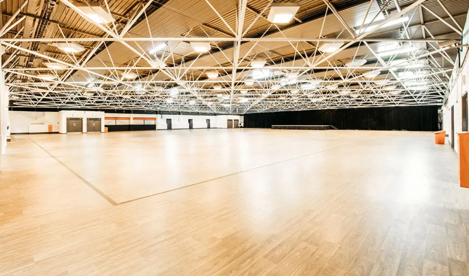 Empty sports hall with wooden floor, white industrial ceiling with exposed steel beams and overhead lighting.