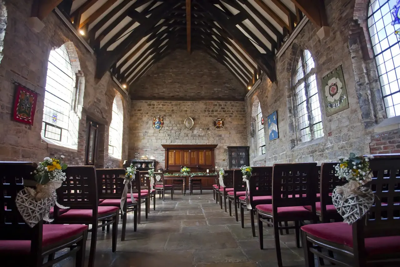 Interior of a historic stone hall with wooden beams, arranged with black chairs with pink cushions, white flower arrangements, and stained-glass windows.