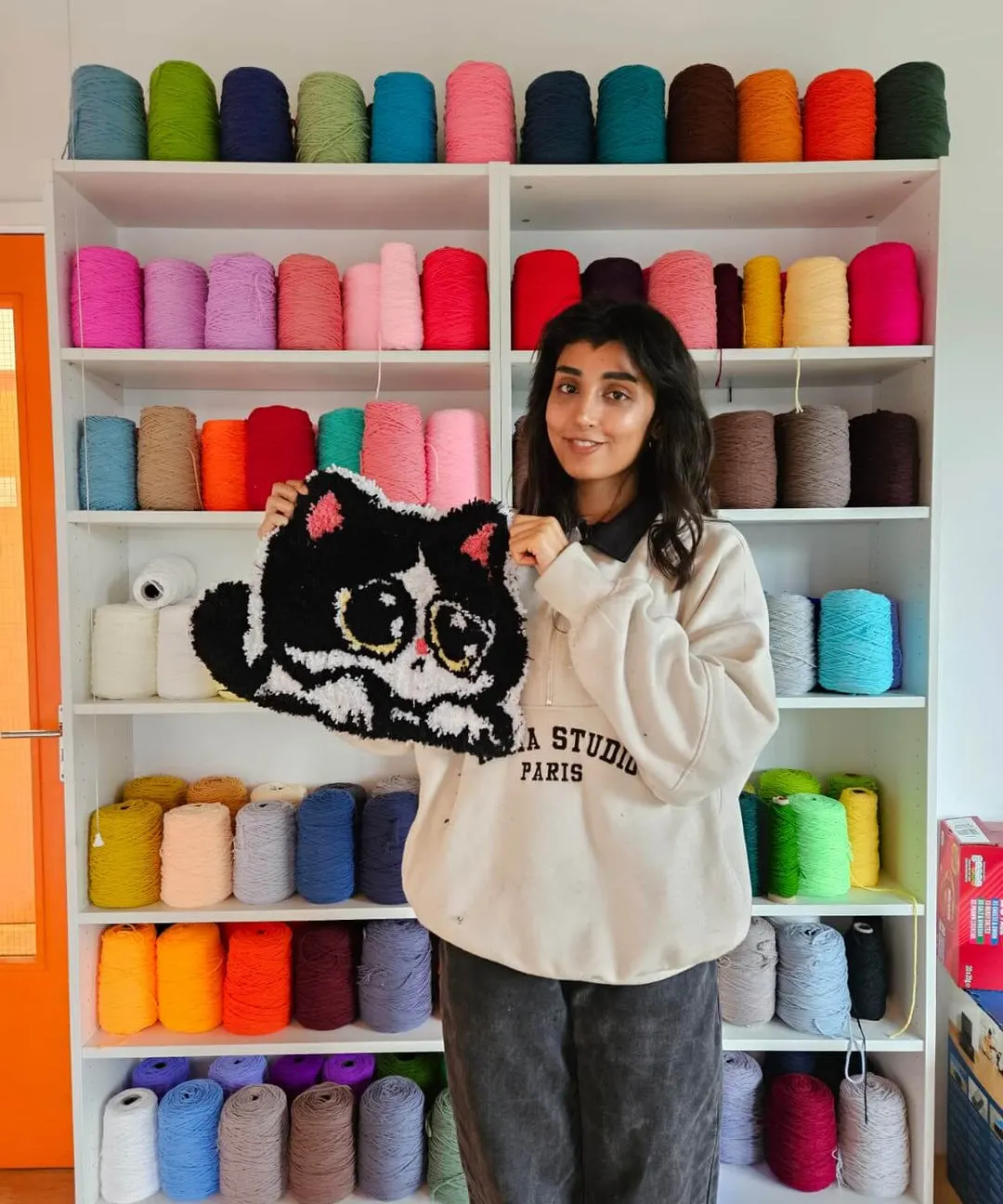 Woman holding a black tufted cat cushion in front of shelves stocked with colourful yarn spools.
