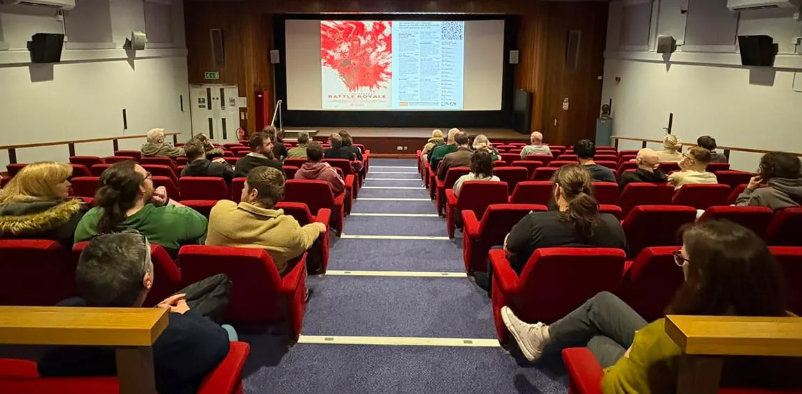 Audience members seated in red cinema seats watching a presentation on a large screen in an auditorium.