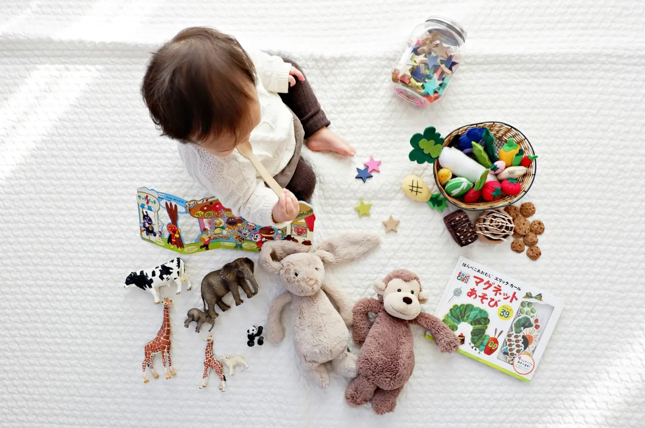 Toddler playing on white surface surrounded by toys including stuffed animals, picture books, toy food and animals.