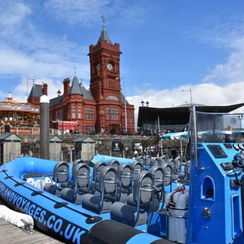 A bright blue ride vehicle with empty seats in the foreground, with Cardiff Castle's red Victorian clock tower and surrounding buildings visible in the background under blue sky.