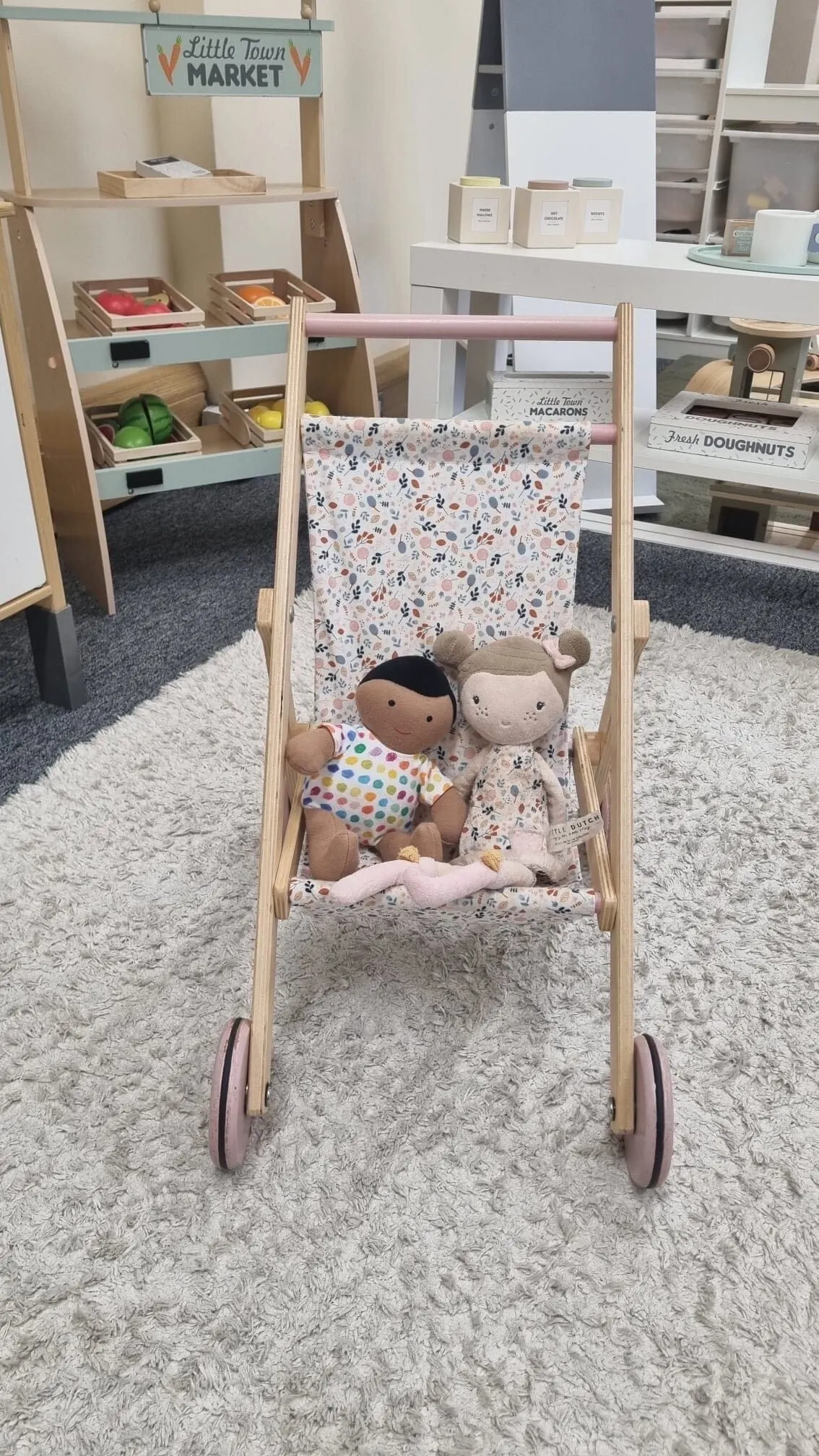 Wooden toy pram with fabric lining containing two soft dolls in a children's play area with market stall displays.