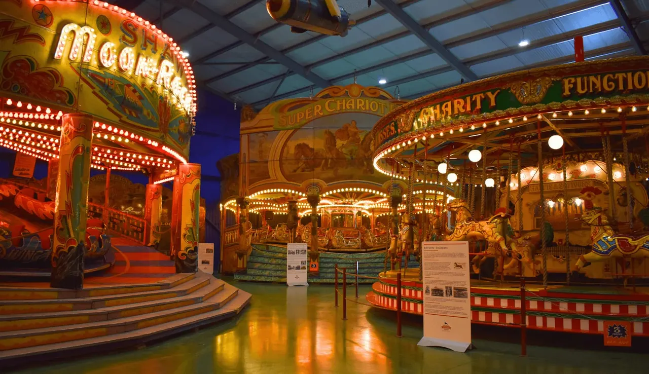 Historic illuminated carousel and fairground rides with hand-painted panels and bulbous lights displayed indoors at a museum.