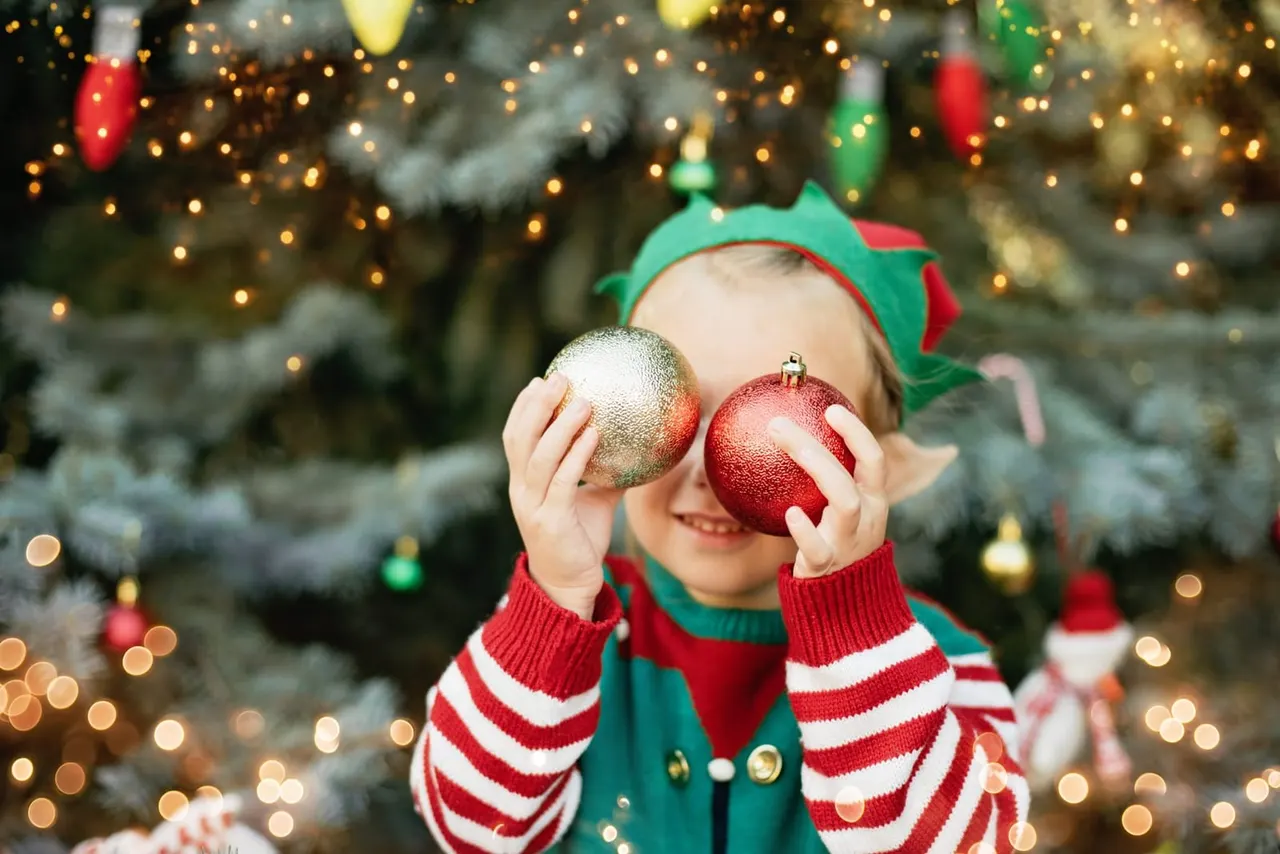 Young child in elf costume holding gold and red baubles in front of decorated Christmas tree with lights.