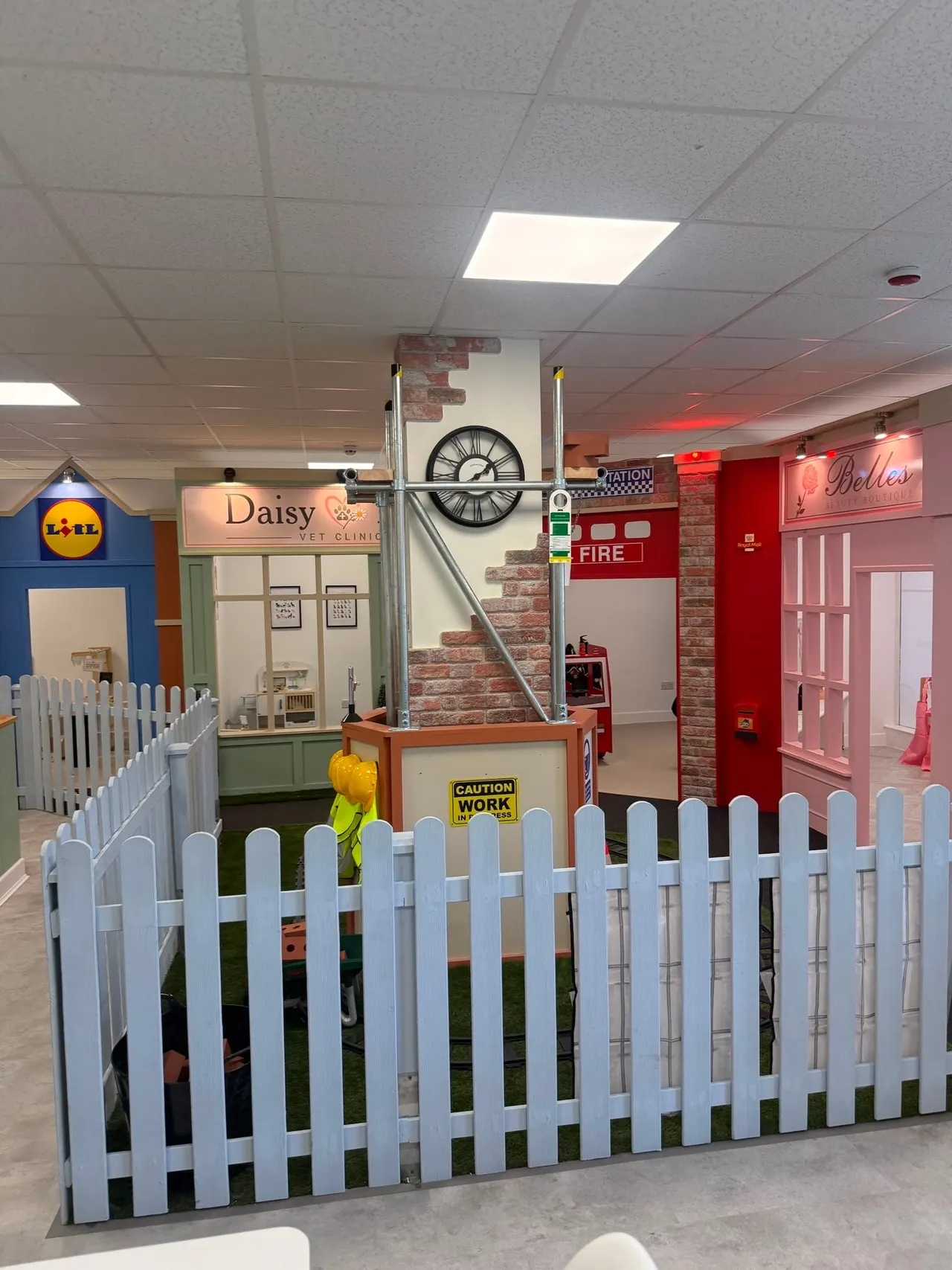 Indoor play area with themed role-play zones including a vet clinic, fire station and shop, separated by white picket fencing.