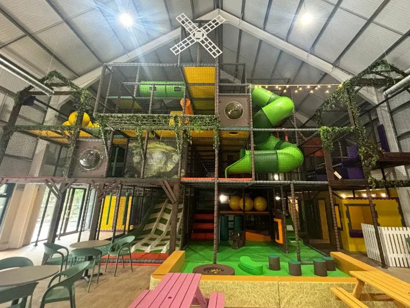 Multi-level indoor play structure with climbing frames, slides, and soft play equipment in a barn-like venue.