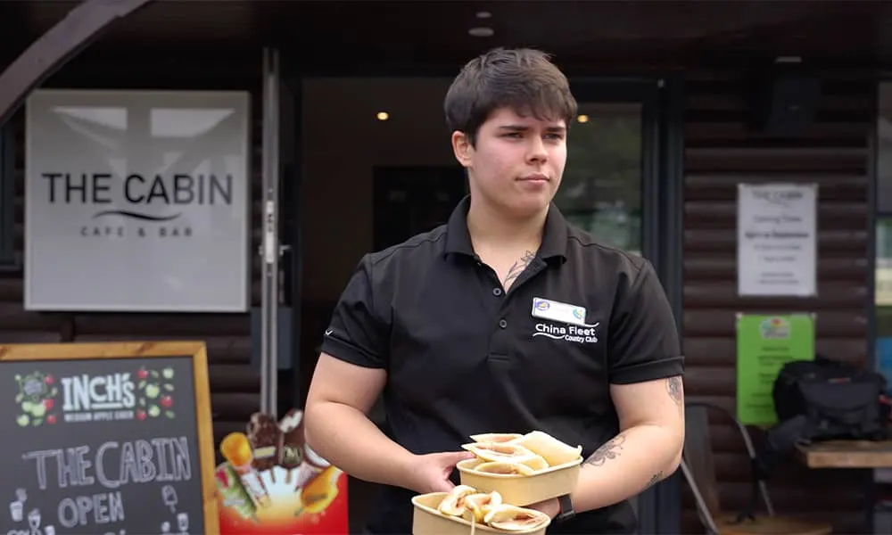 Young staff member holding two plates of chips outside The Cabin cafe and bar entrance.