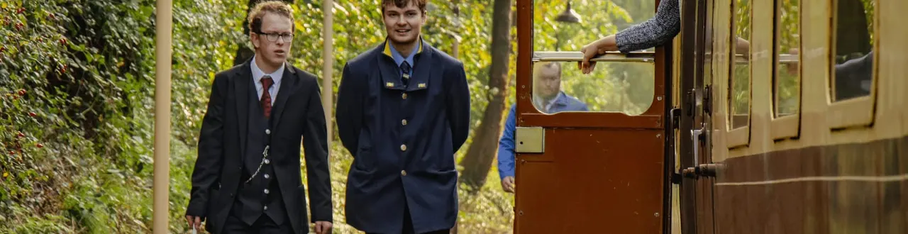 Two men in smart uniforms walk alongside a vintage railway carriage surrounded by green foliage.