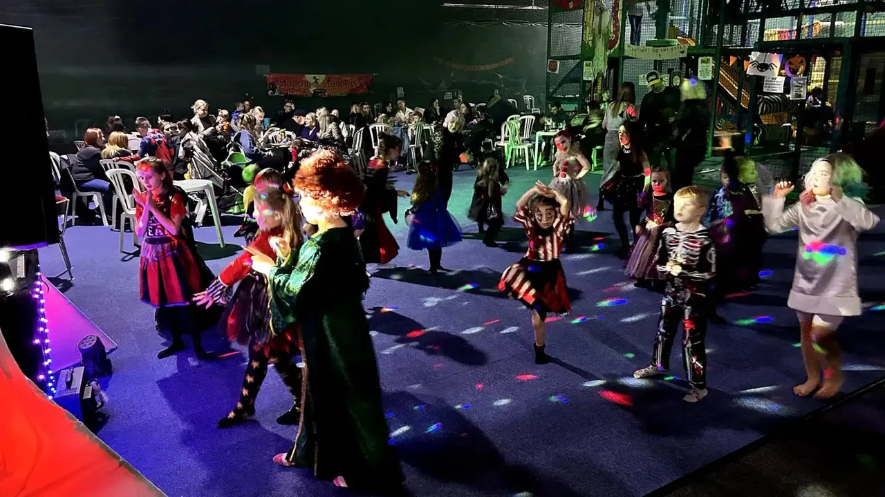 Children in Halloween costumes dancing and playing on a lit indoor stage with seated spectators watching from the sides.