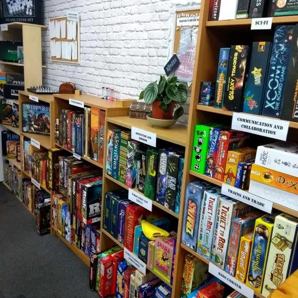 Wooden shelves displaying a large collection of colourful board games and tabletop games, with white brick wall and potted plant.
