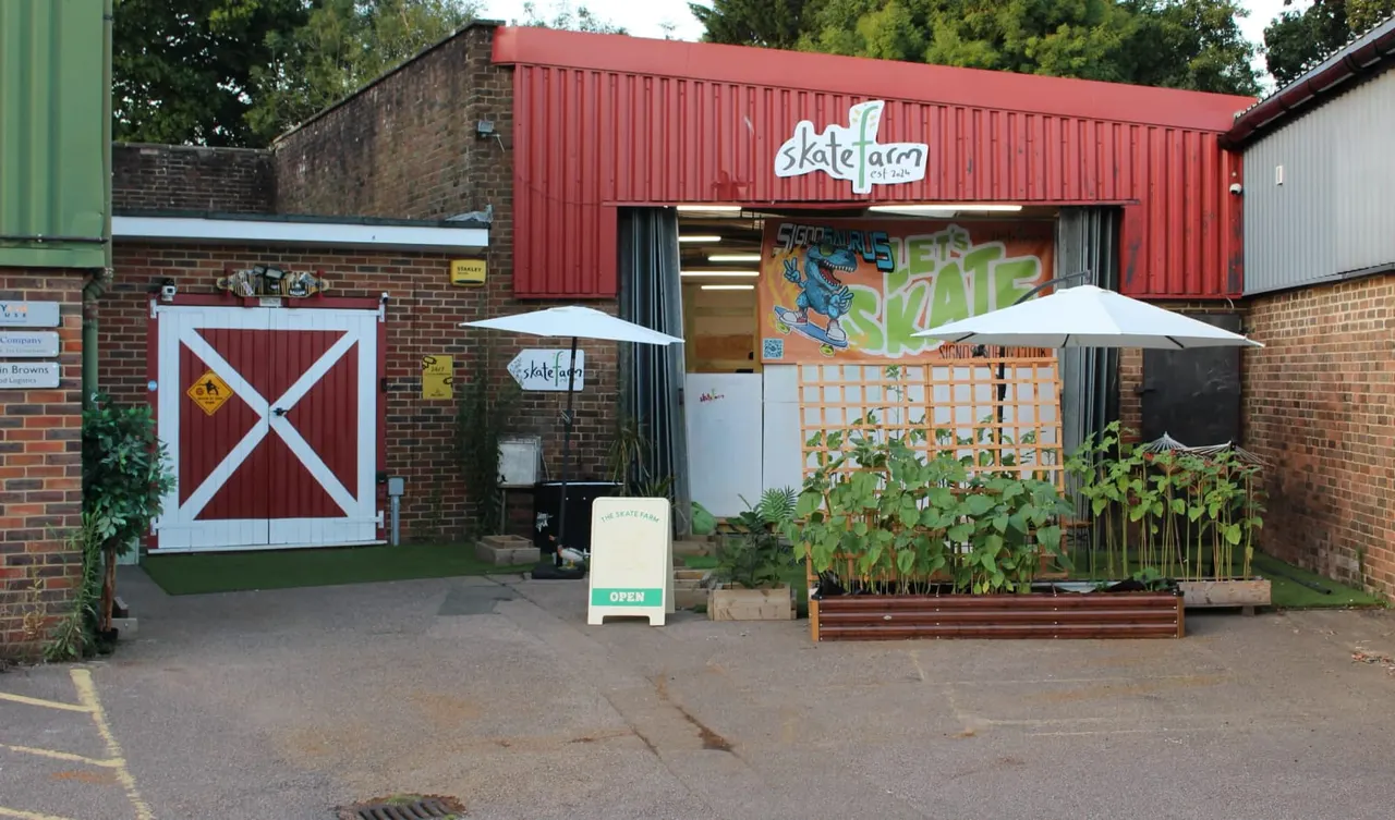Exterior of a skateboarding venue with red corrugated metal building, outdoor seating under umbrellas, and planted areas.