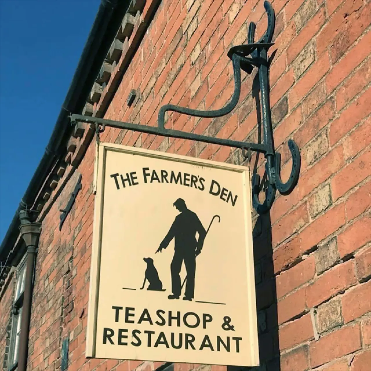 A cream-coloured pub sign for The Farmer's Den Teashop & Restaurant mounted on a wrought-iron bracket against red brick.