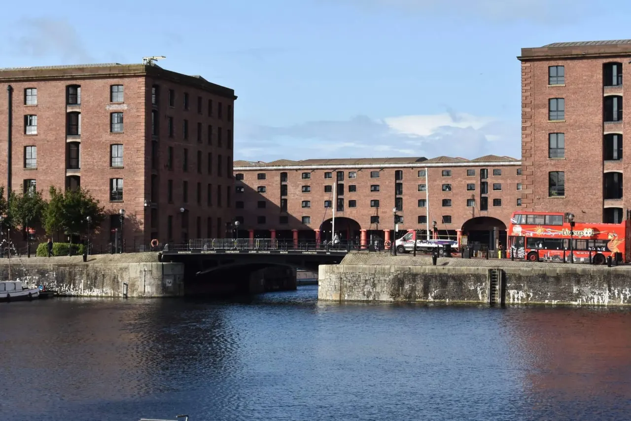 Red tour bus parked at a waterfront dock surrounded by Victorian brick warehouses and a stone bridge over the canal.