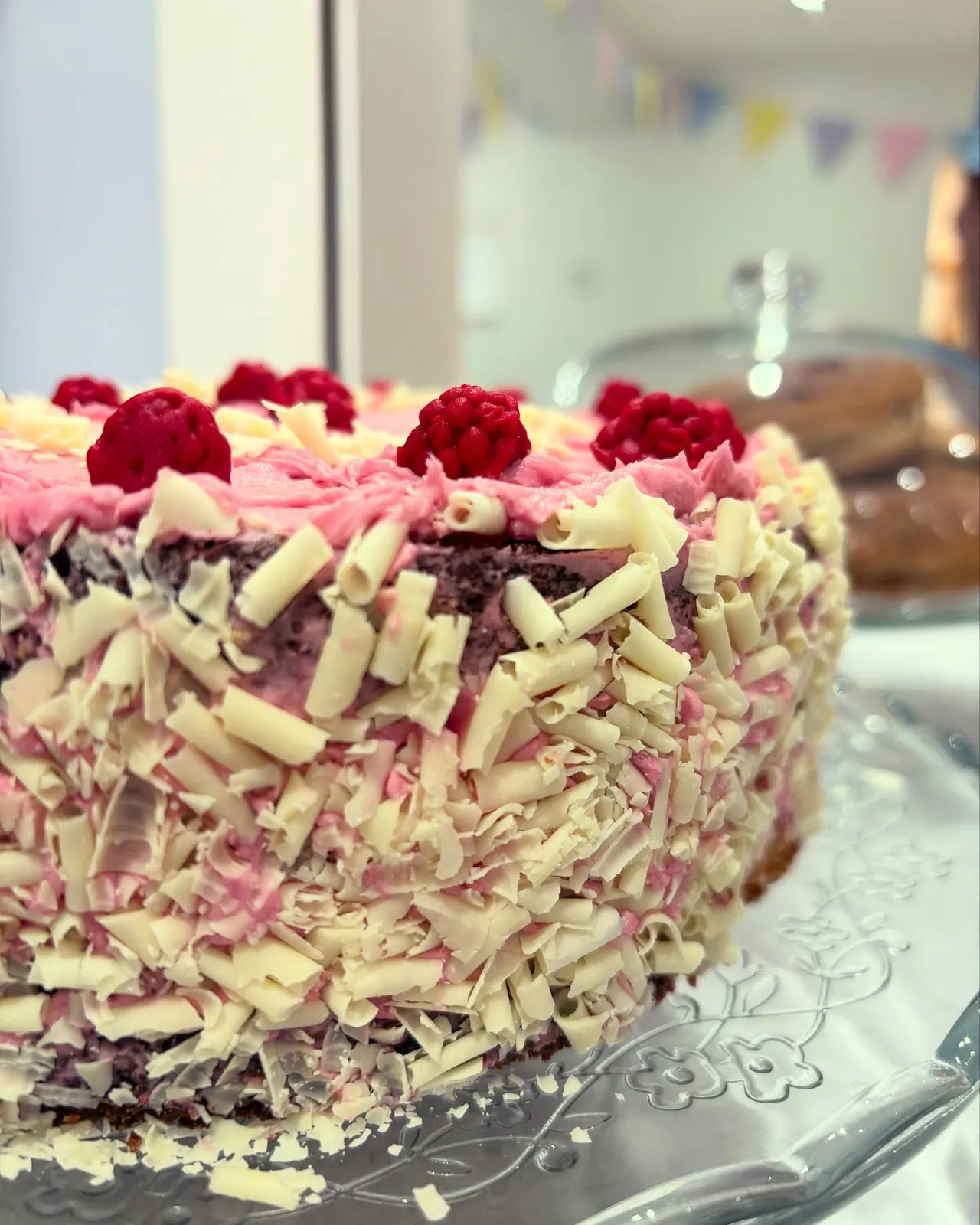 Decorated cake with pink frosting, white chocolate shavings, and fresh raspberries on top, displayed on a silver cake server.