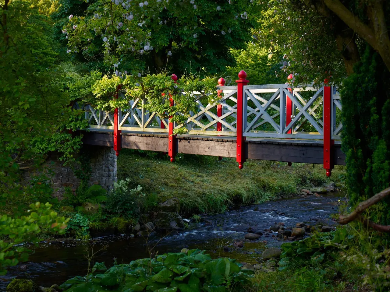 Decorative wooden bridge with red railings and white lattice panels spanning a small stream, surrounded by lush green foliage and flowering climbing plants.