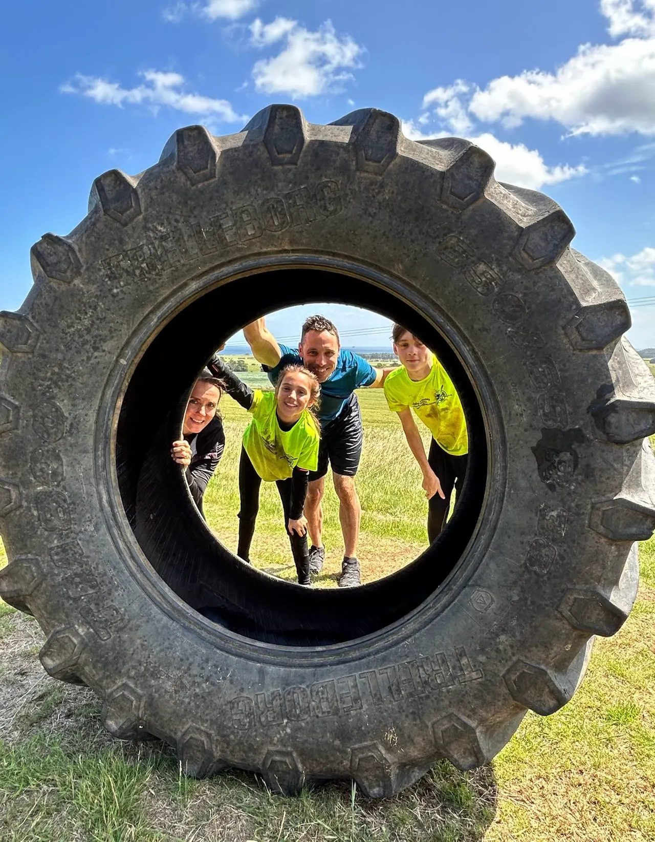 Five people in bright yellow and blue tops posing together through a large tractor tyre on a grassy field.