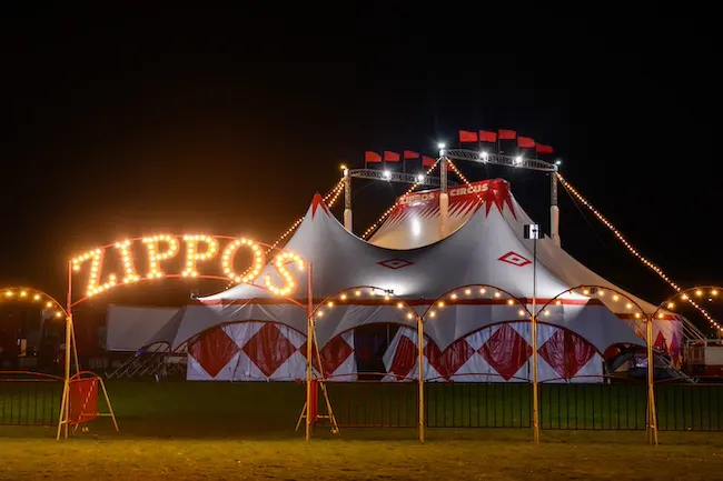 Illuminated circus tent with red and white striped design and "ZIPPOS" sign lit up at night.