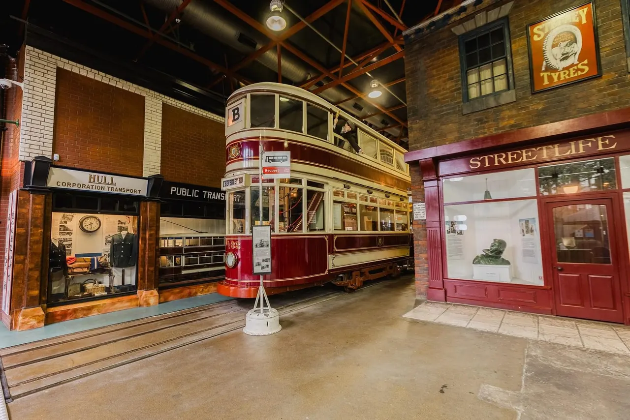 A vintage red and cream double-decker tram on display indoors, flanked by period shop fronts including Hull Corporation Transport and Streetlife storefronts.