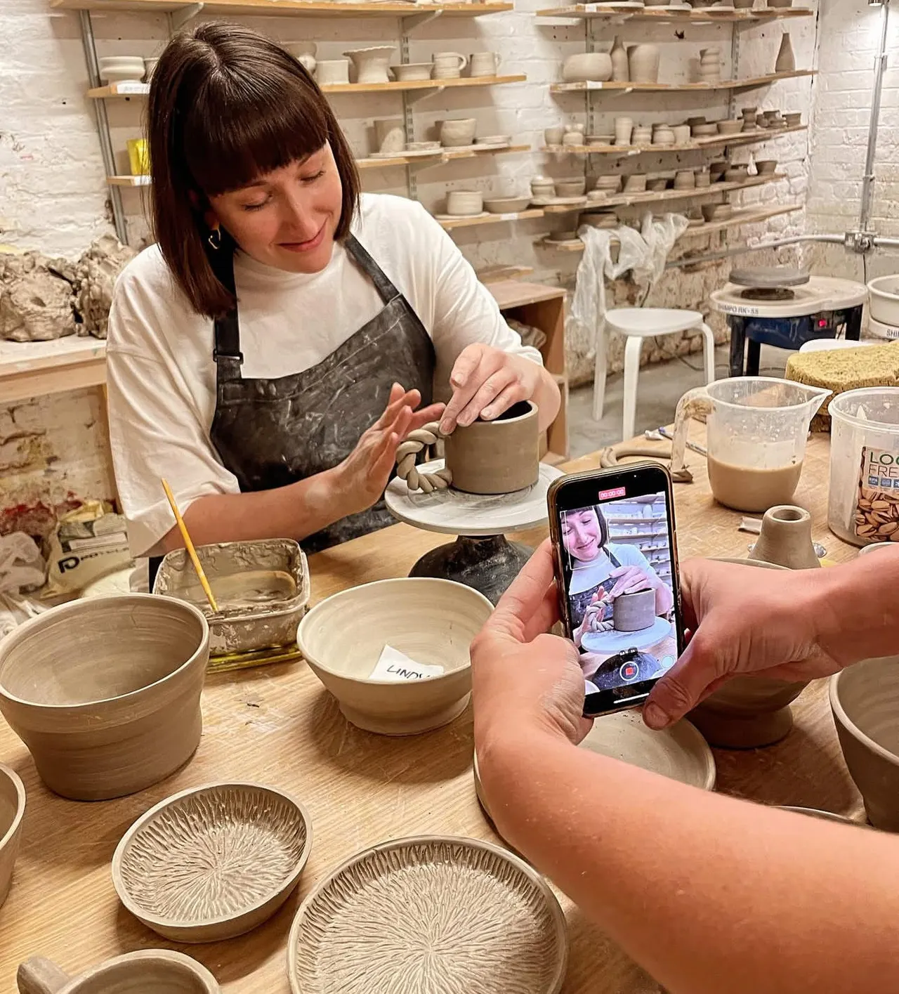A potter in an apron works at a pottery wheel whilst someone photographs her with a phone in a studio lined with shelves of ceramic pieces.