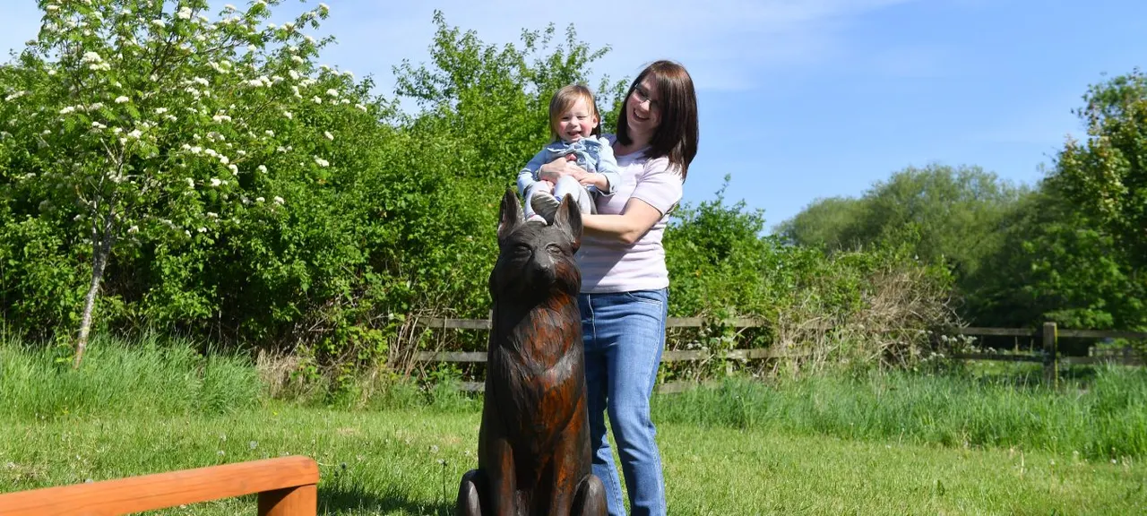 Woman holding a young child next to a large wooden carved animal sculpture in a sunny park setting.
