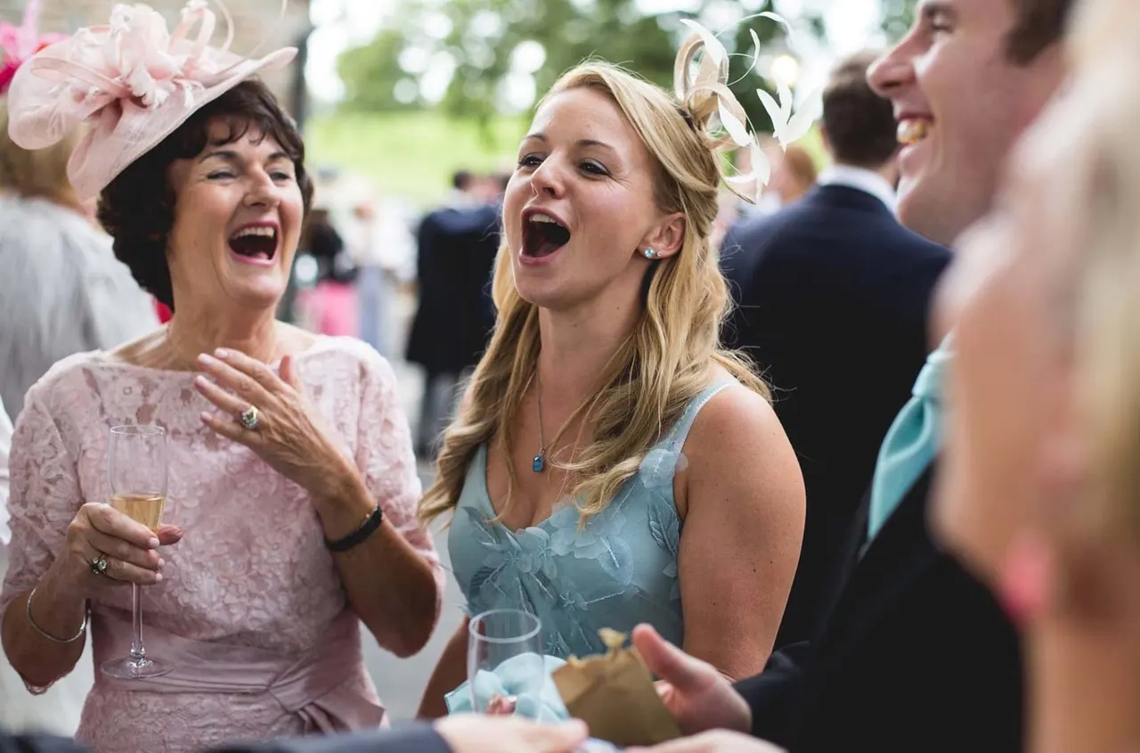 Well-dressed guests laughing and holding drinks at an outdoor garden party, wearing fascinators and formal attire.