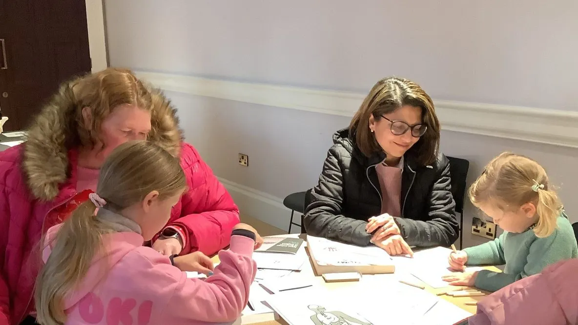 An adult and three children sit at a table doing a drawing or art activity together indoors.