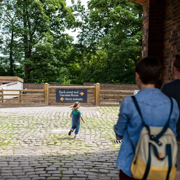 A young child runs across a paved courtyard towards a directional sign, with an adult in a blue jacket watching nearby.