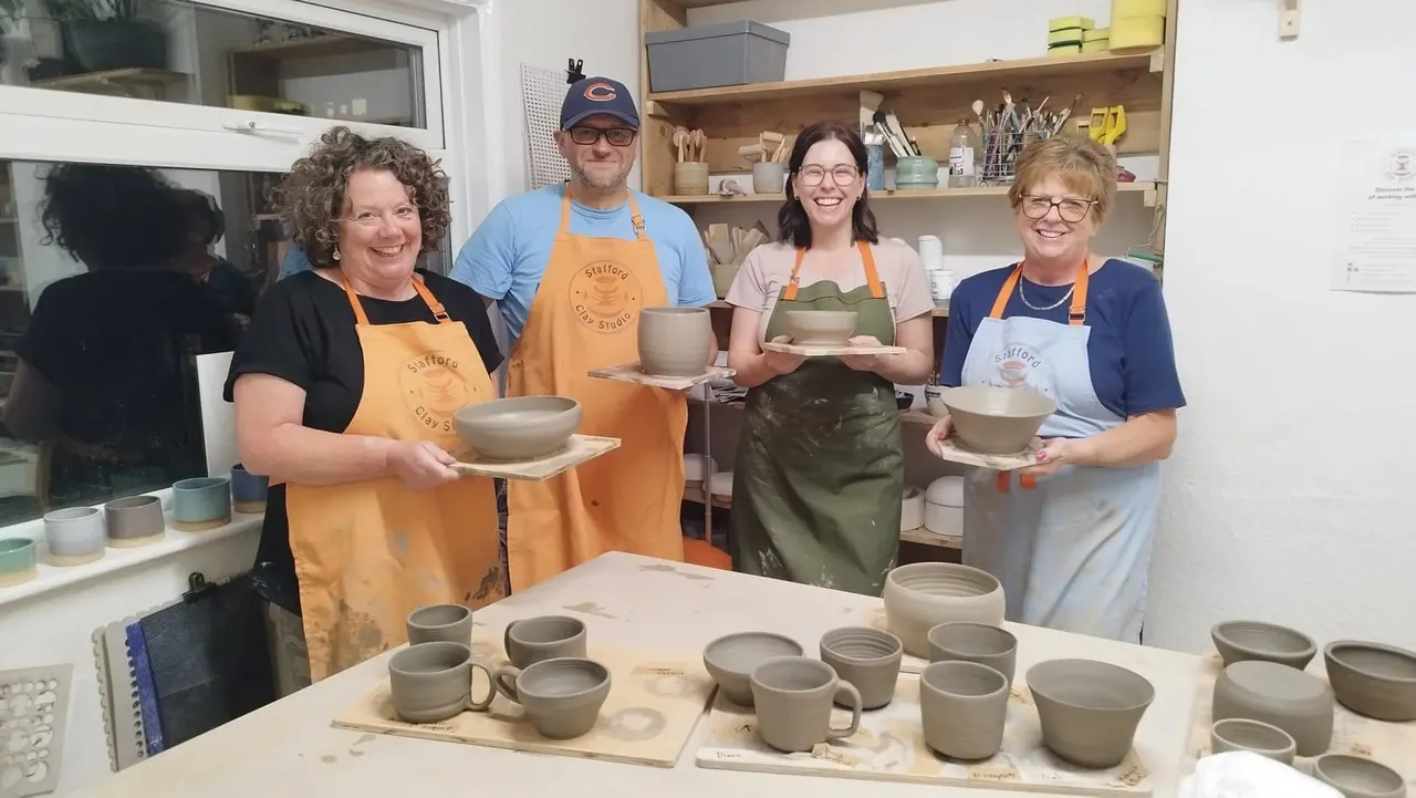 Four people wearing aprons in a pottery studio display handmade ceramic bowls and mugs they have created.