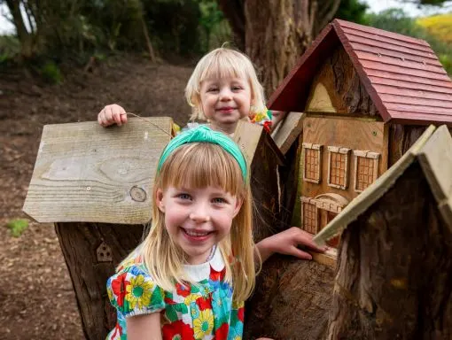 Two young children smiling at wooden play structures in a woodland setting.