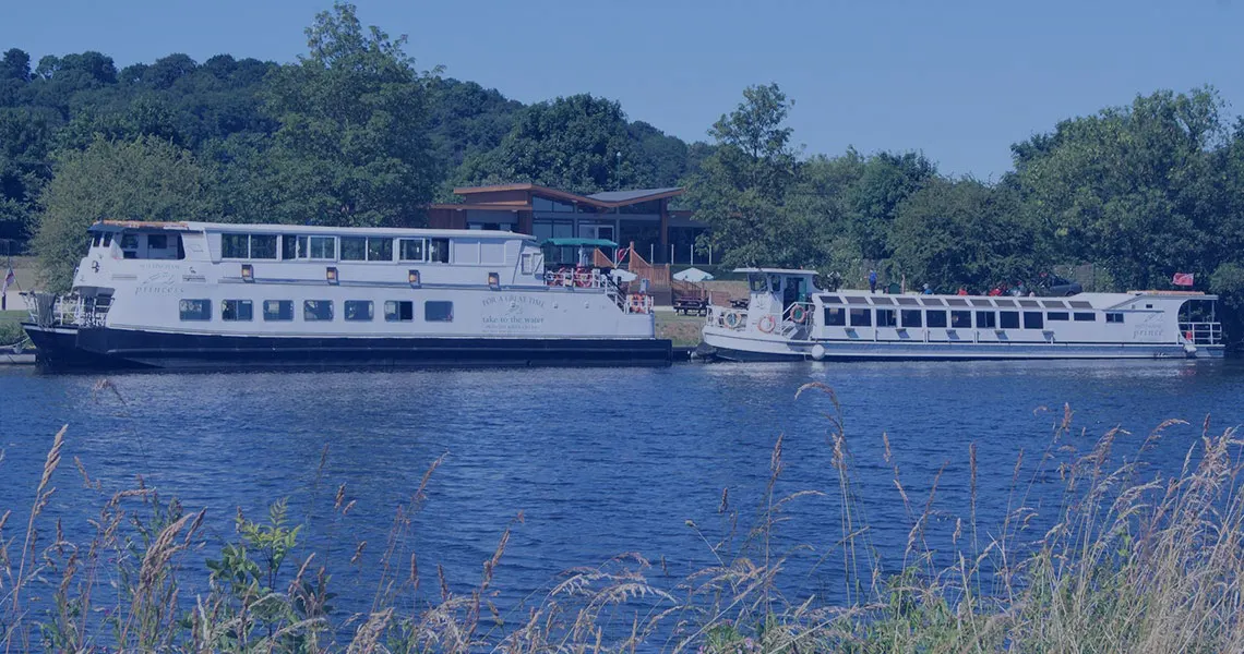 Two river cruise vessels moored on blue water with trees lining the banks and reeds in the foreground.