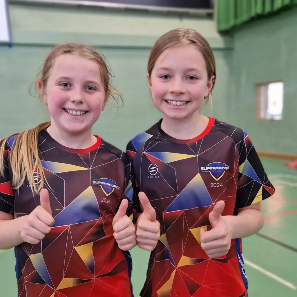 Two young girls in matching team sports uniforms giving thumbs up in an indoor sports hall.