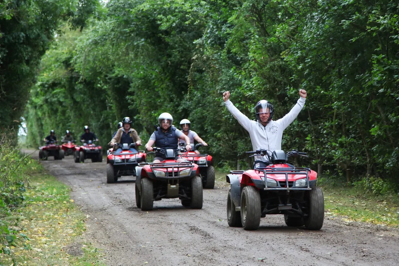 Group of people riding red quad bikes on a tree-lined dirt track, with the front rider celebrating with arms raised.
