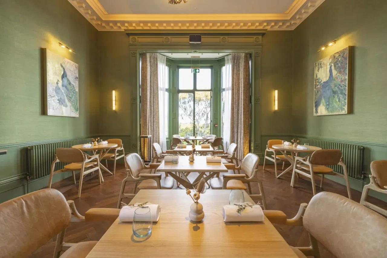 Elegant dining room with sage-green walls, wooden tables set with napkins, and peacock artwork, opening onto gardens through tall green-framed doors.