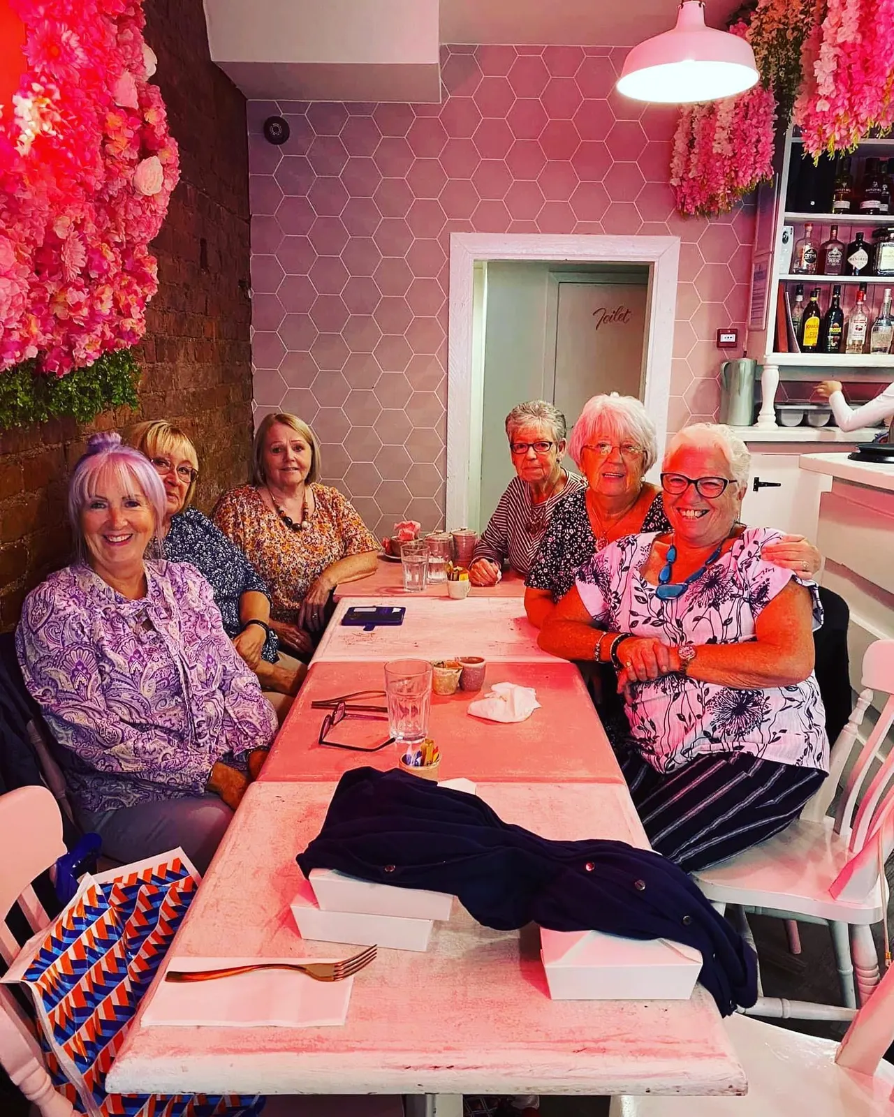 A group of six older women sitting at a pink table in a brightly decorated pink dining room with floral wall installations.