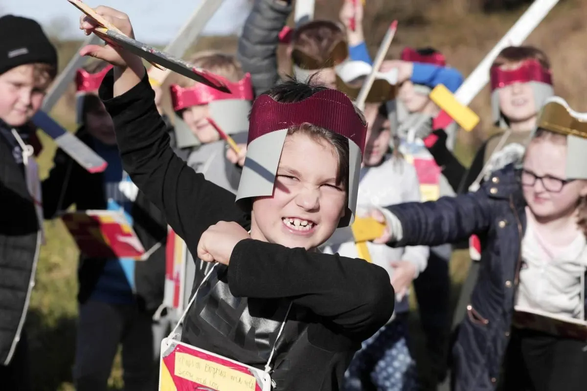 A child in a maroon and white hat waves a stick enthusiastically whilst surrounded by other children holding coloured flags outdoors.