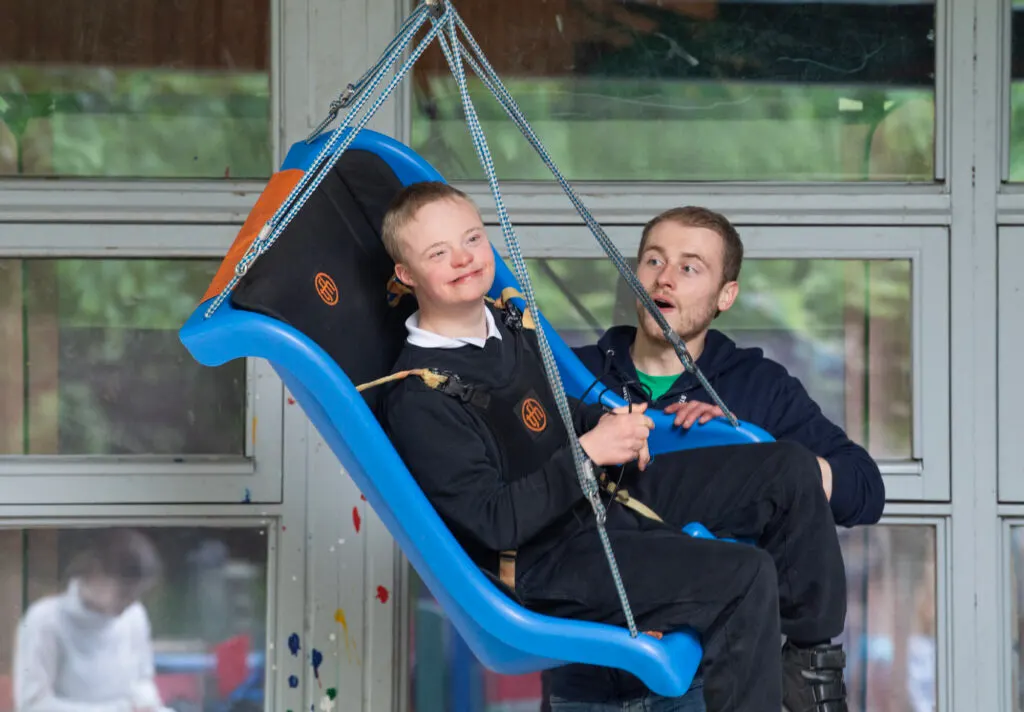 Young child and adult on an indoor swing ride against glass doors with garden views.