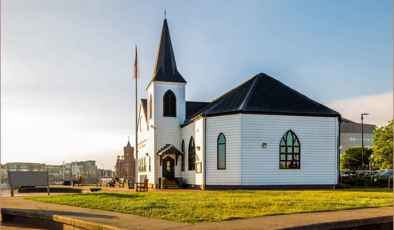 White wooden church building with black spire and gold trim, surrounded by grass and paving, beside modern buildings on a waterfront.
